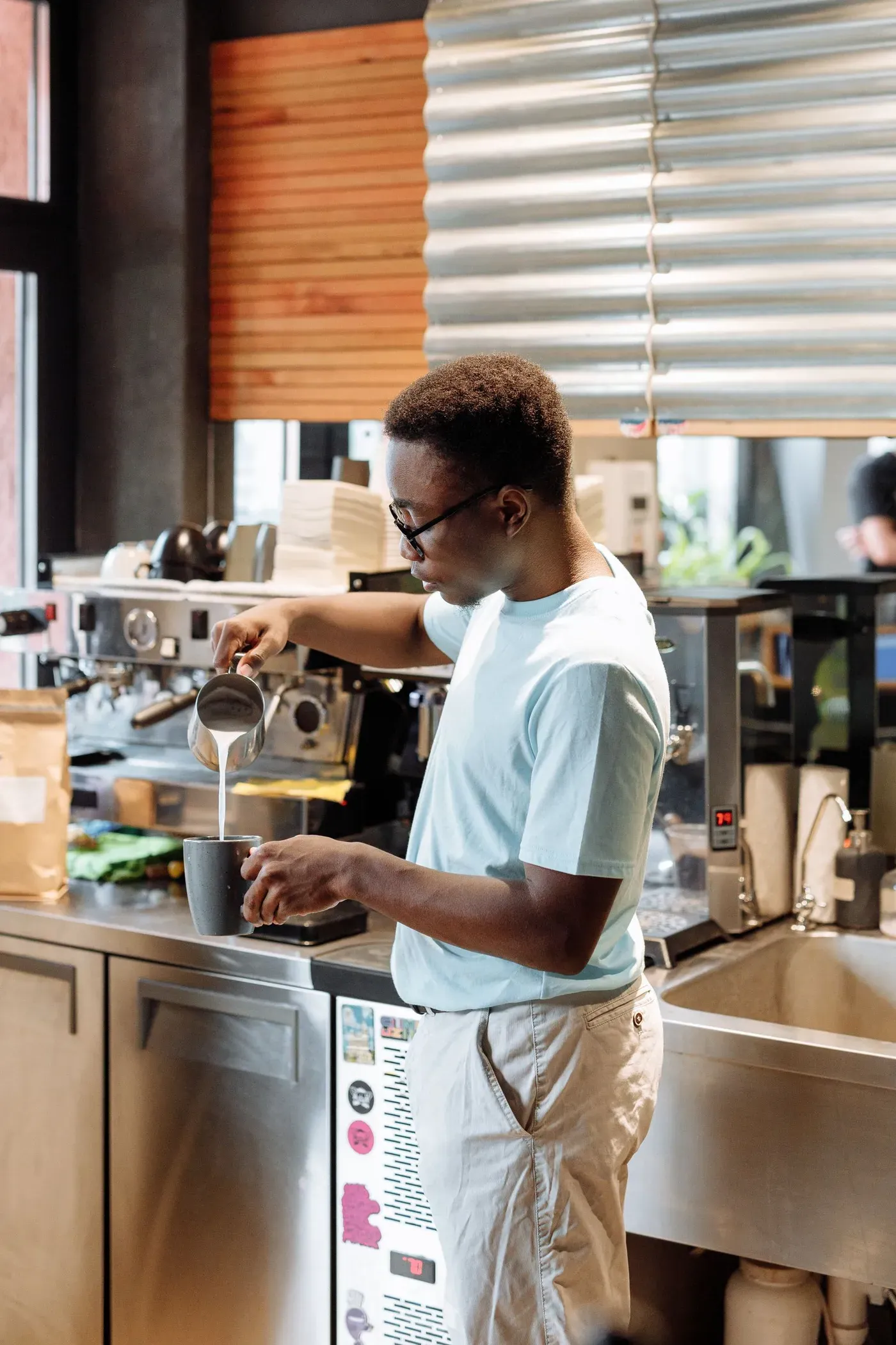 A barista in a small cafe pouring steamed milk into a cup at the espresso counter.