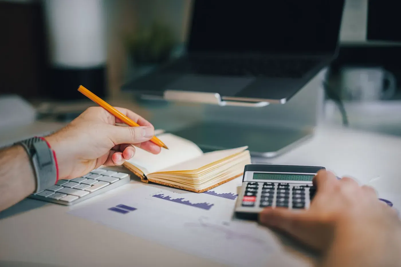 Hands working with a calculator, a notebook and printed charts beside a laptop.