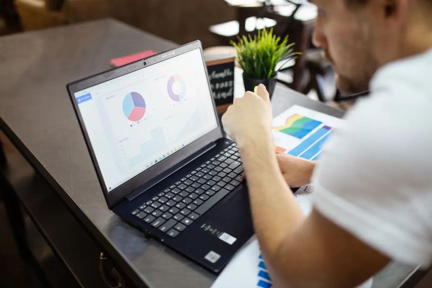 A man reviewing pie charts and bar charts on a laptop with printed reports beside him.