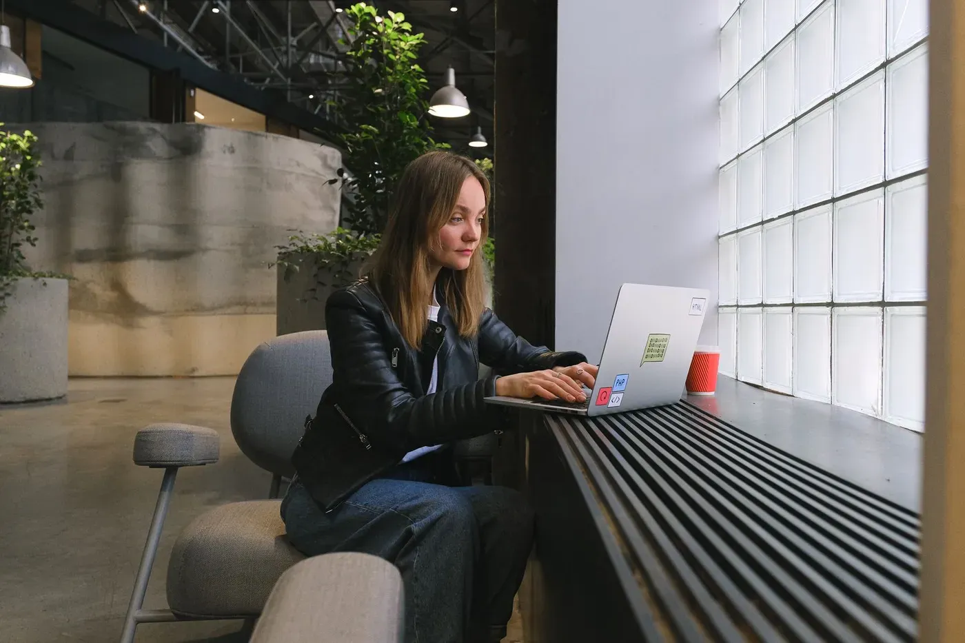 A web designer working on a laptop at a window desk in a bright office.
