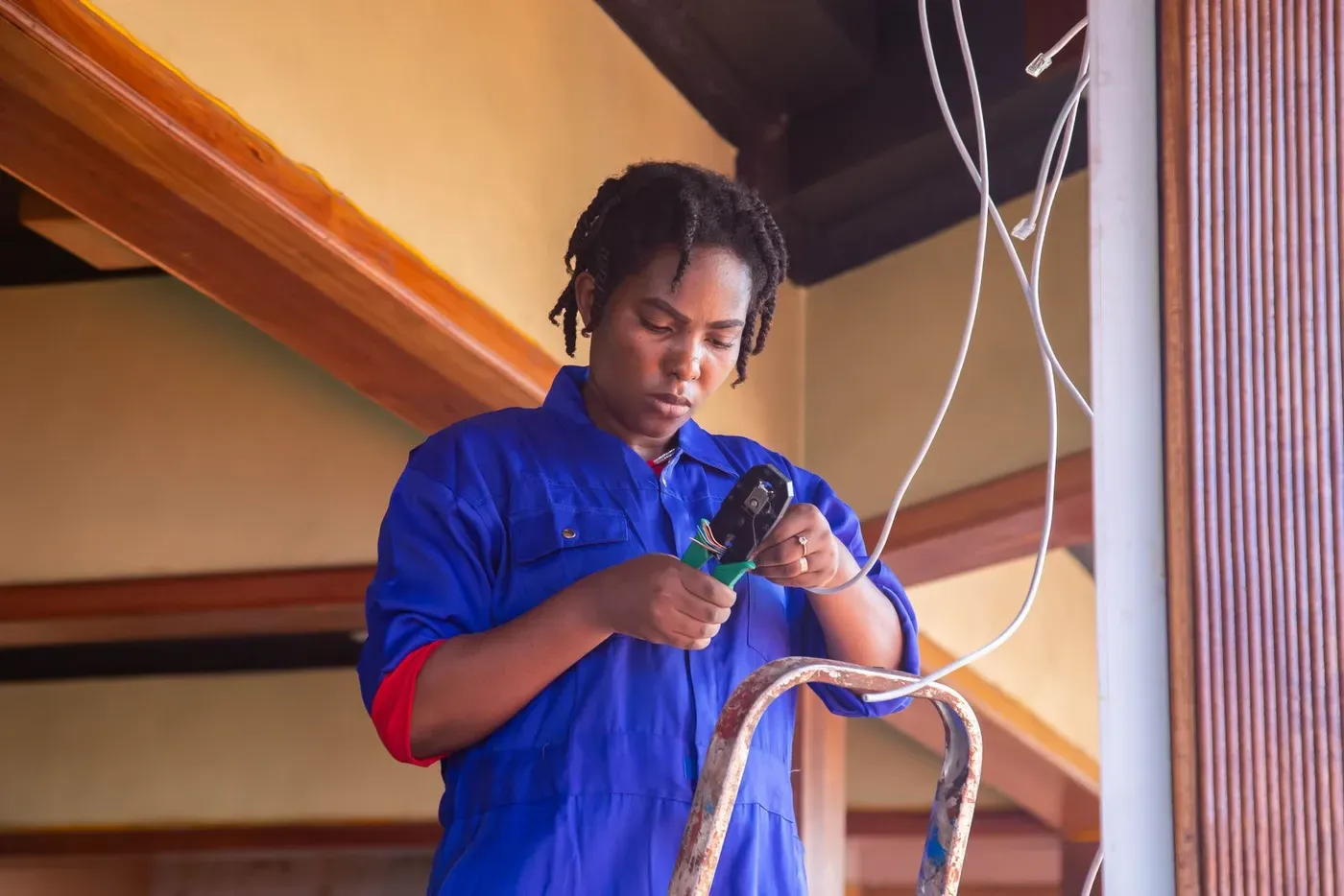 An electrician in blue overalls crimping a cable on a ladder.