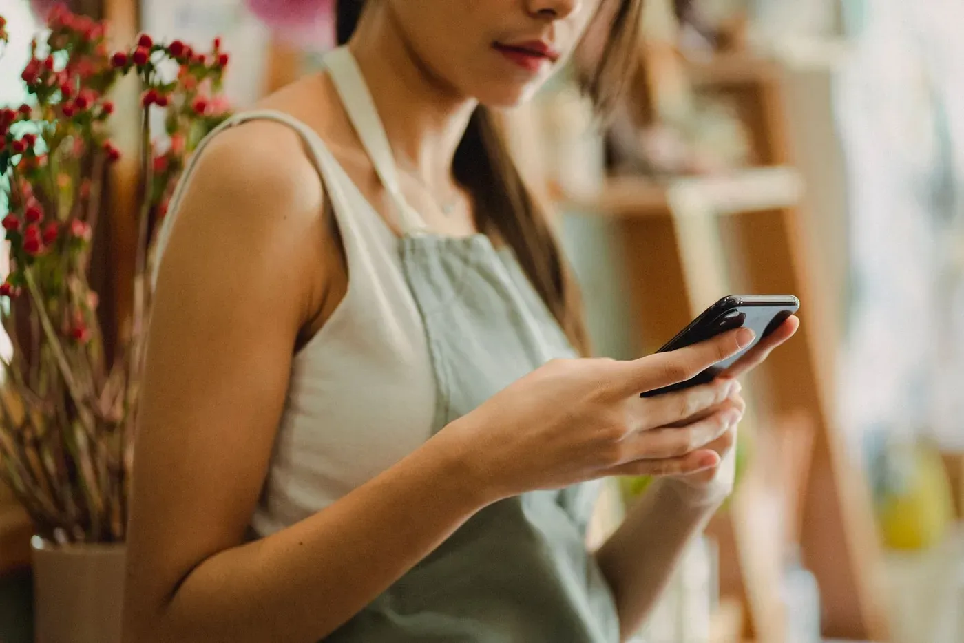 A florist in an apron checking a phone beside stems of red berries in her shop.