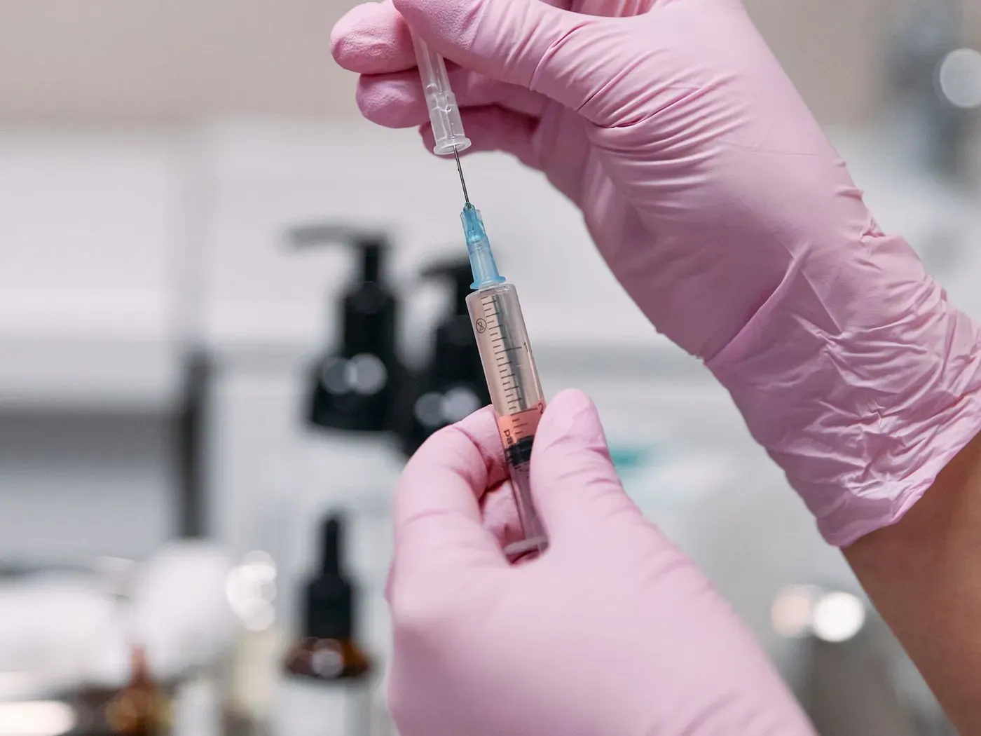 Hands in pink gloves holding a syringe in a clinic, with bottles out of focus behind.