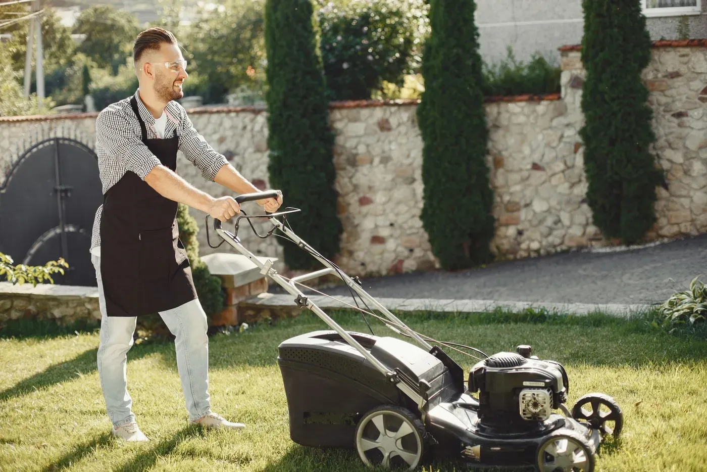 A man in safety glasses and an apron pushing a lawn mower across a garden.