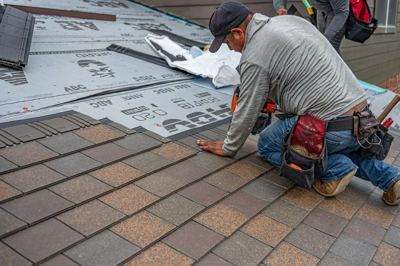 A roofer kneeling on a roof and fixing shingles in place with a nail gun.