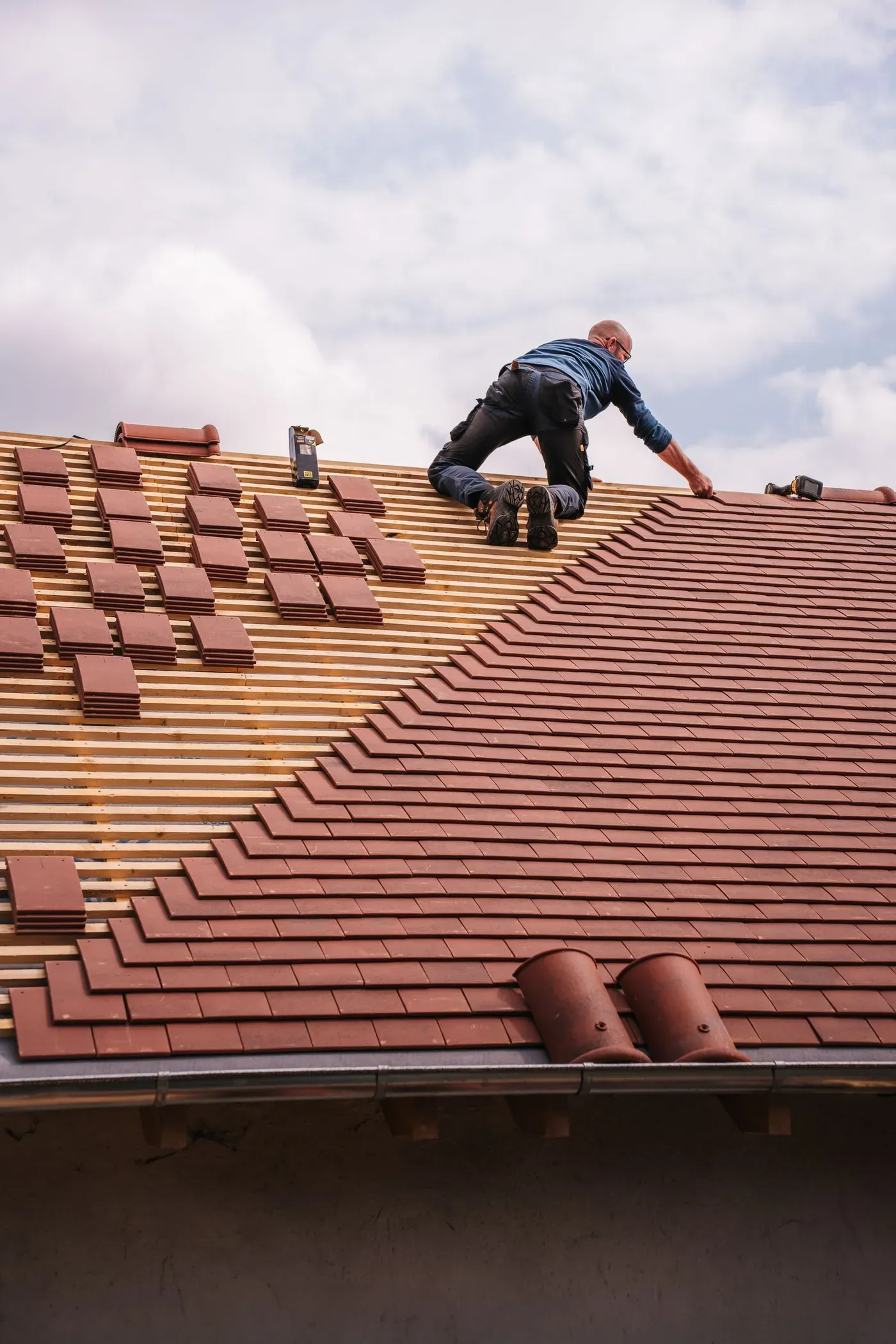 A roofer kneeling near the ridge of a pitched roof, laying red clay tiles over wooden battens.