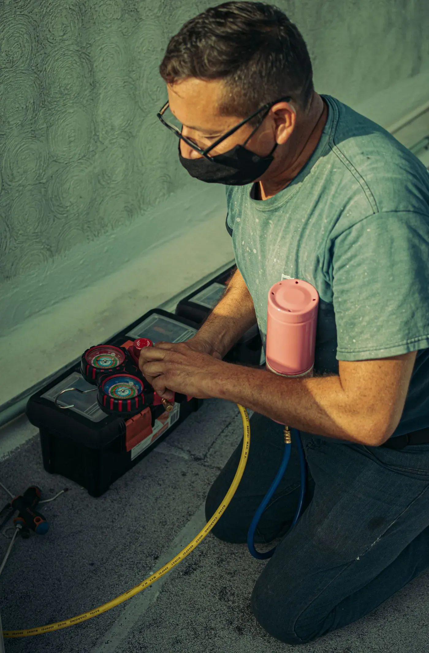 A kneeling technician in a face mask adjusting a set of refrigerant manifold gauges.