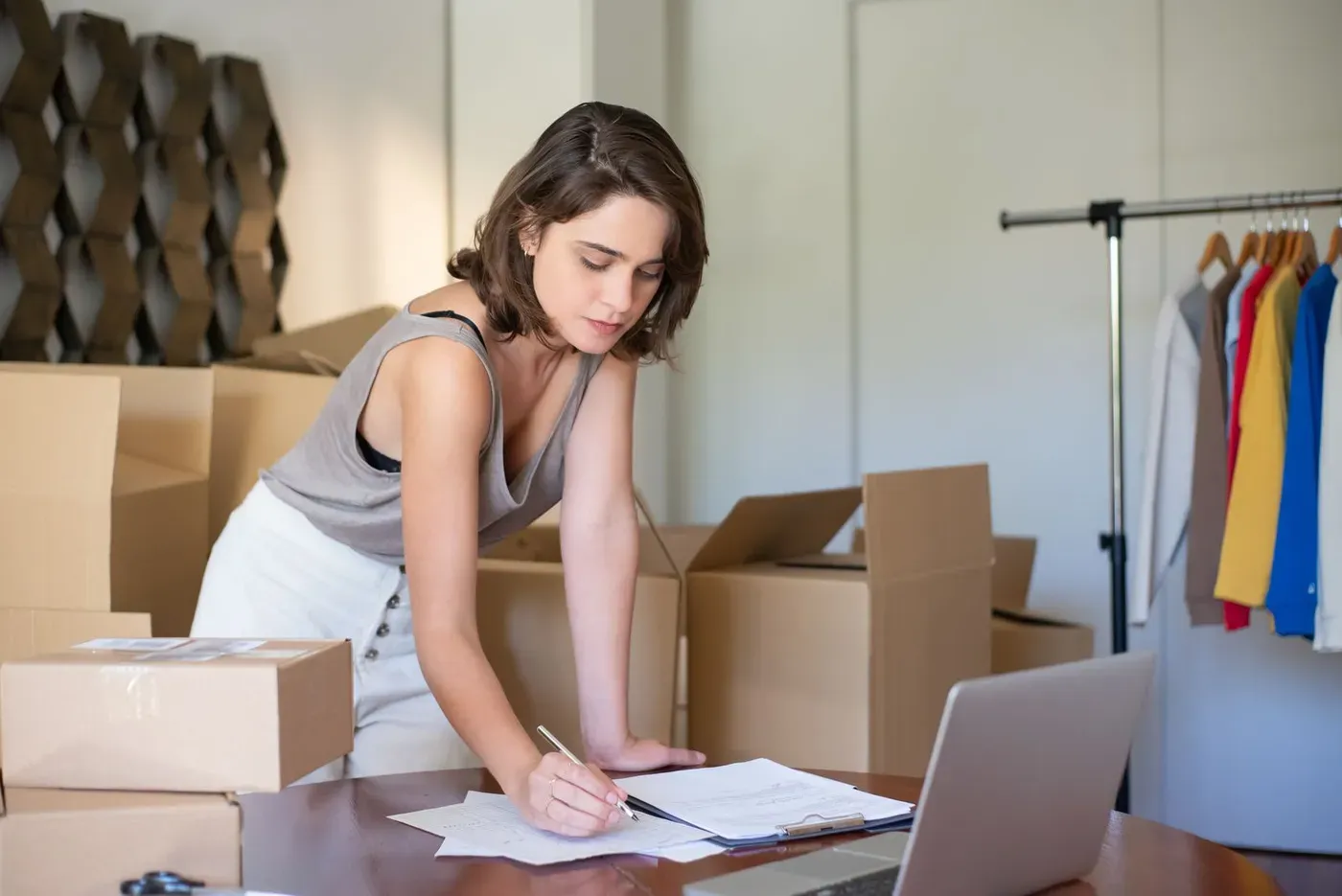 A shop owner writing notes beside a laptop, with packing boxes and a clothes rail behind her.