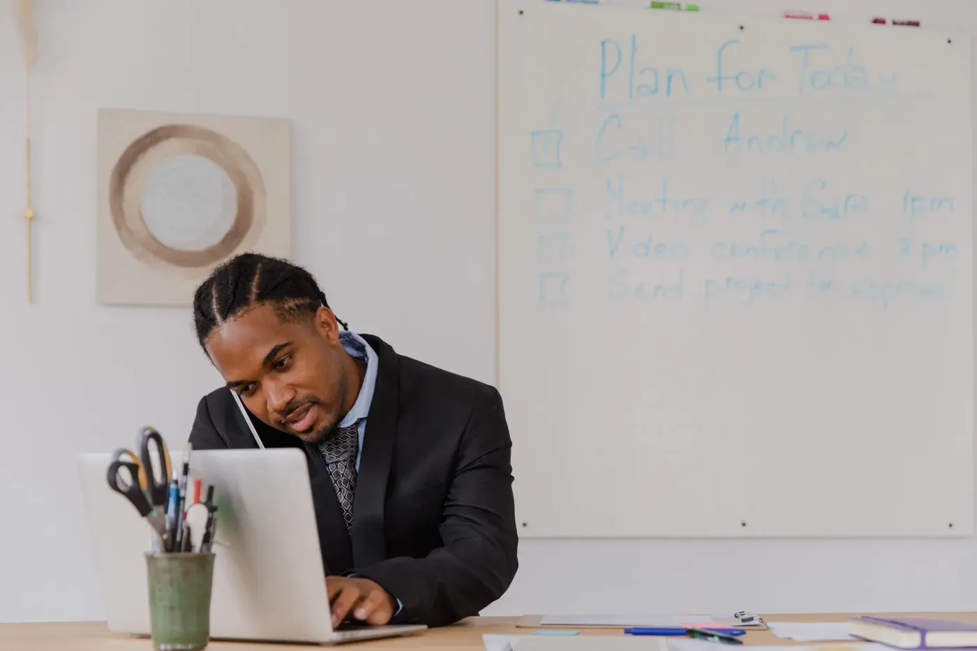 A man in a suit on a phone call while typing on a laptop at a white desk.