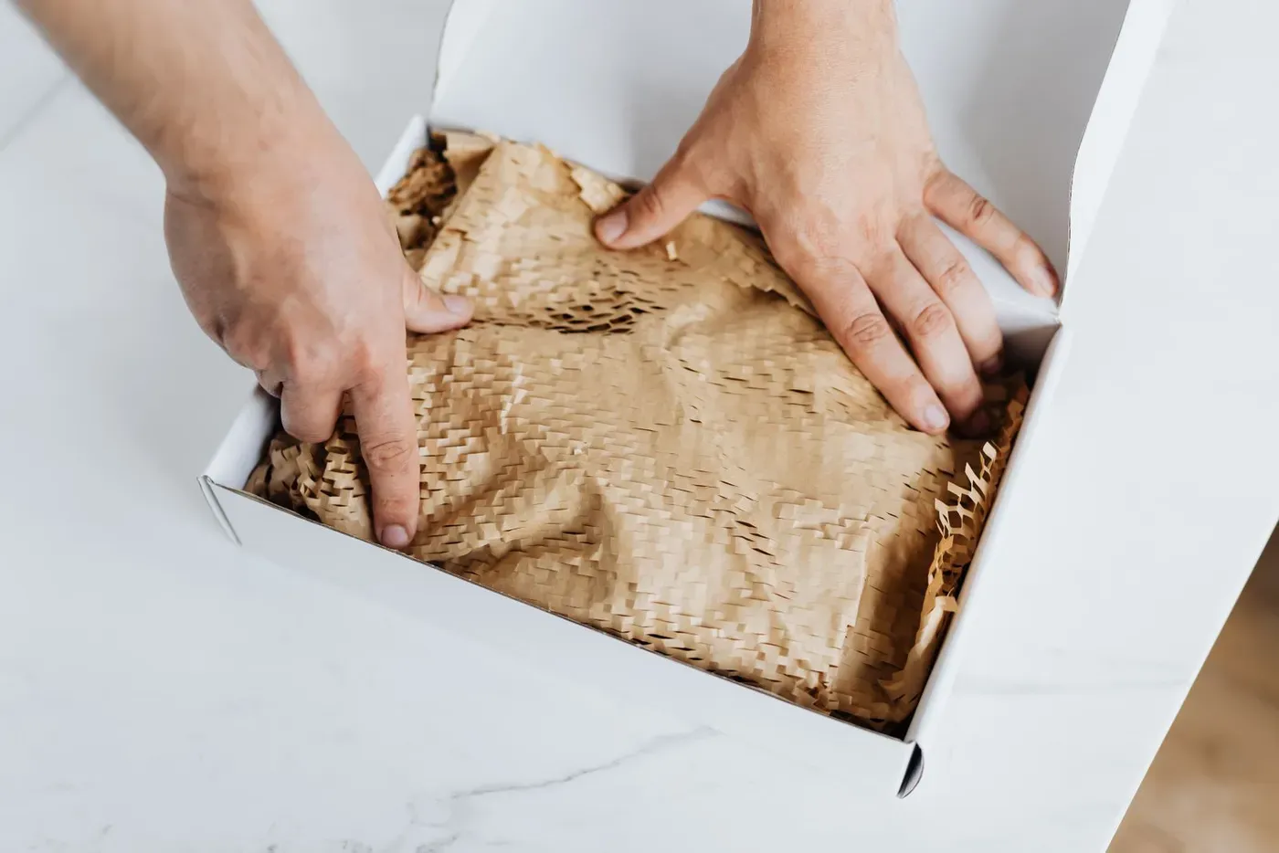Two hands pressing paper padding into a white cardboard shipping box.