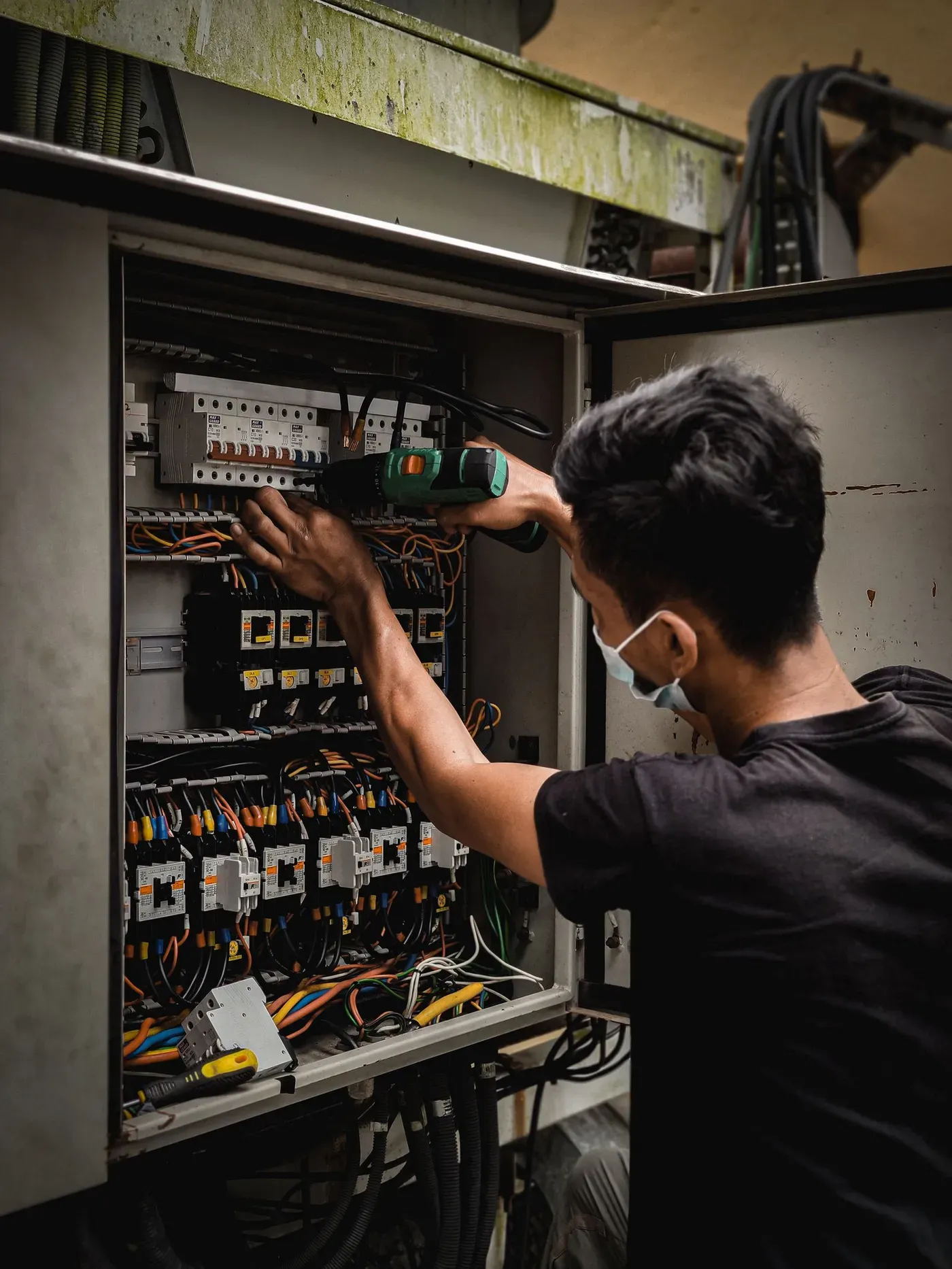 An electrician in a face mask using a cordless drill inside a large open breaker panel.