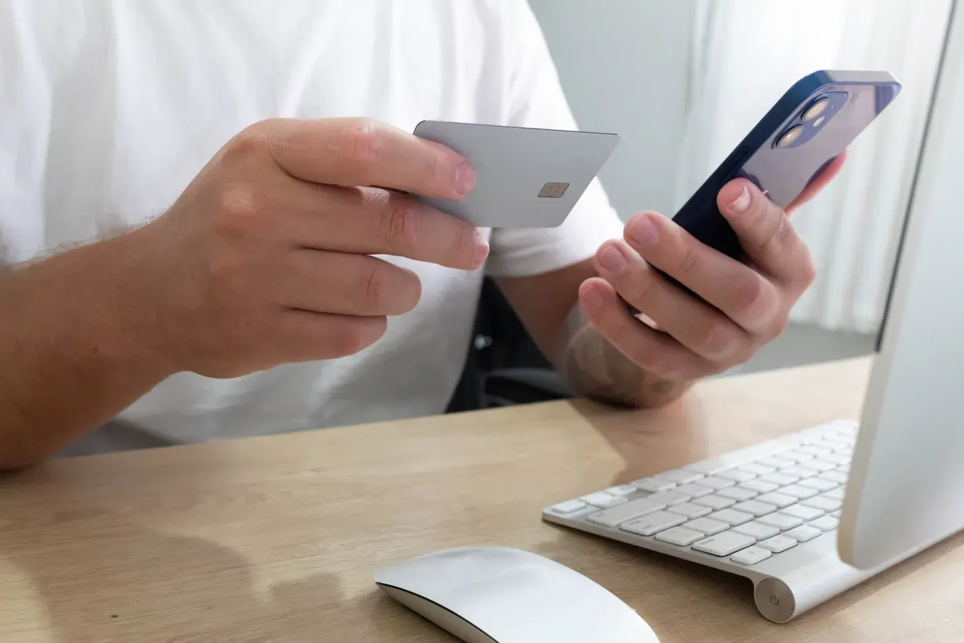 A hand holding a blank payment card beside a phone at a desk with a keyboard.