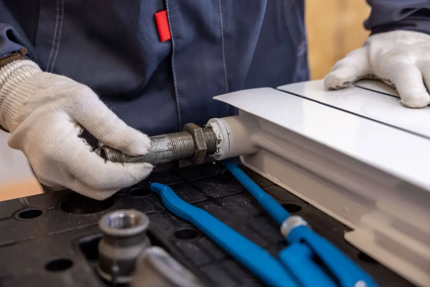 A plumber in work gloves fitting a threaded pipe to a radiator.
