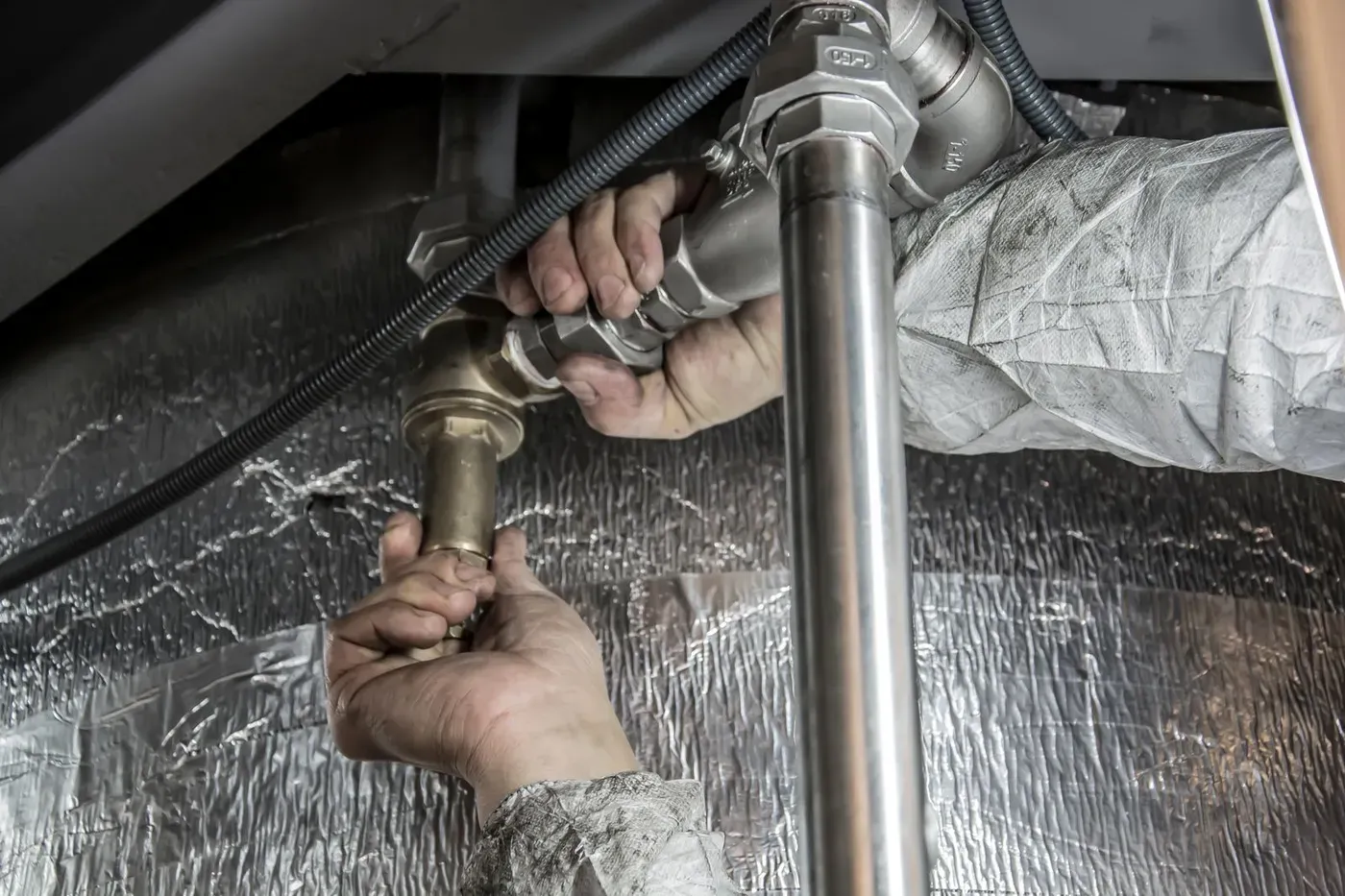 A plumber's hands tightening a brass fitting on pipes under a sink.