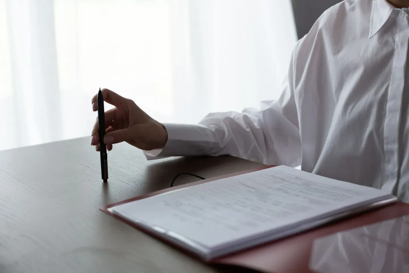 A person holding a pen beside an open folder of printed pages on a desk.