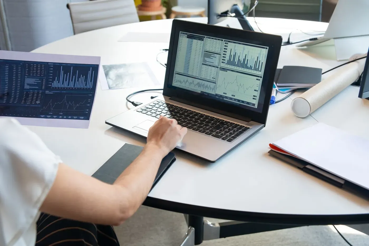 A person working on a laptop showing bar and line charts, with a printed report beside it.