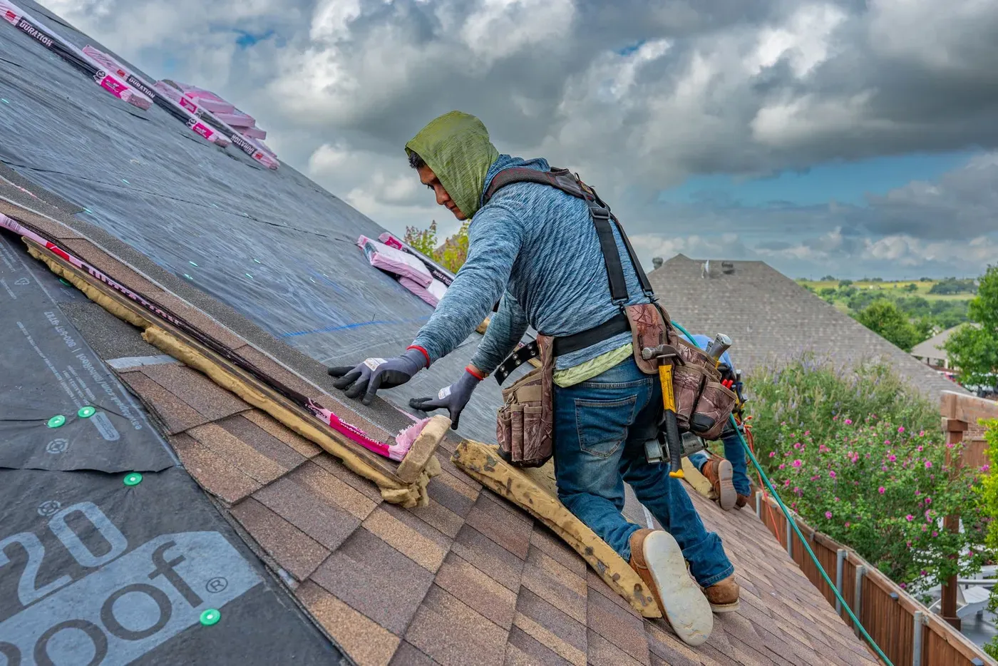 A roofer in a tool belt laying shingles on a steep roof under a cloudy sky.