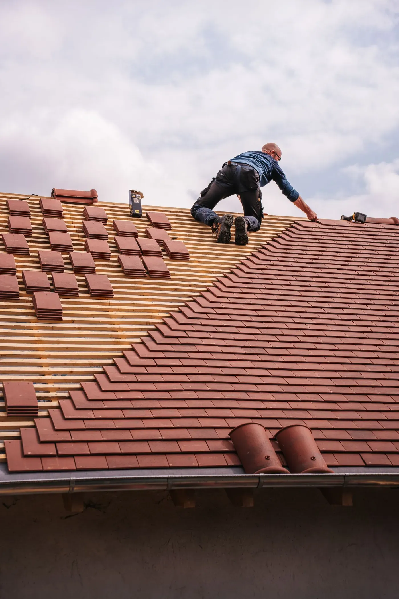 A roofer kneeling on a half-finished clay-tile roof, laying the next rows of tiles.