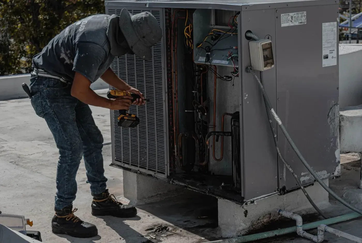 A technician with a cordless drill servicing an open air conditioning unit on a flat roof.