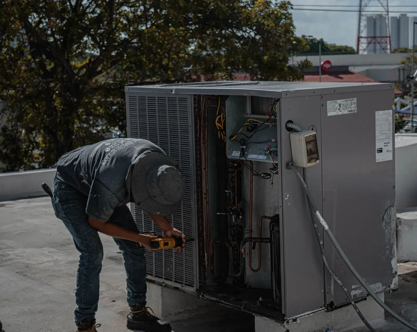 A technician using a cordless drill on an open air conditioning unit on a flat roof.
