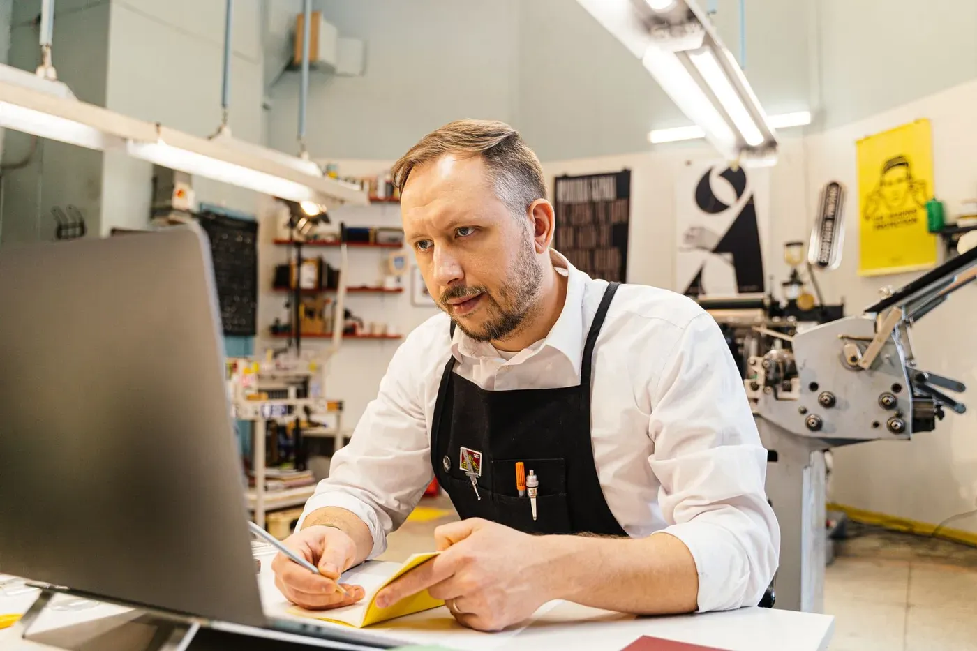 A print shop owner in an apron working at a laptop, making notes beside his printing press.