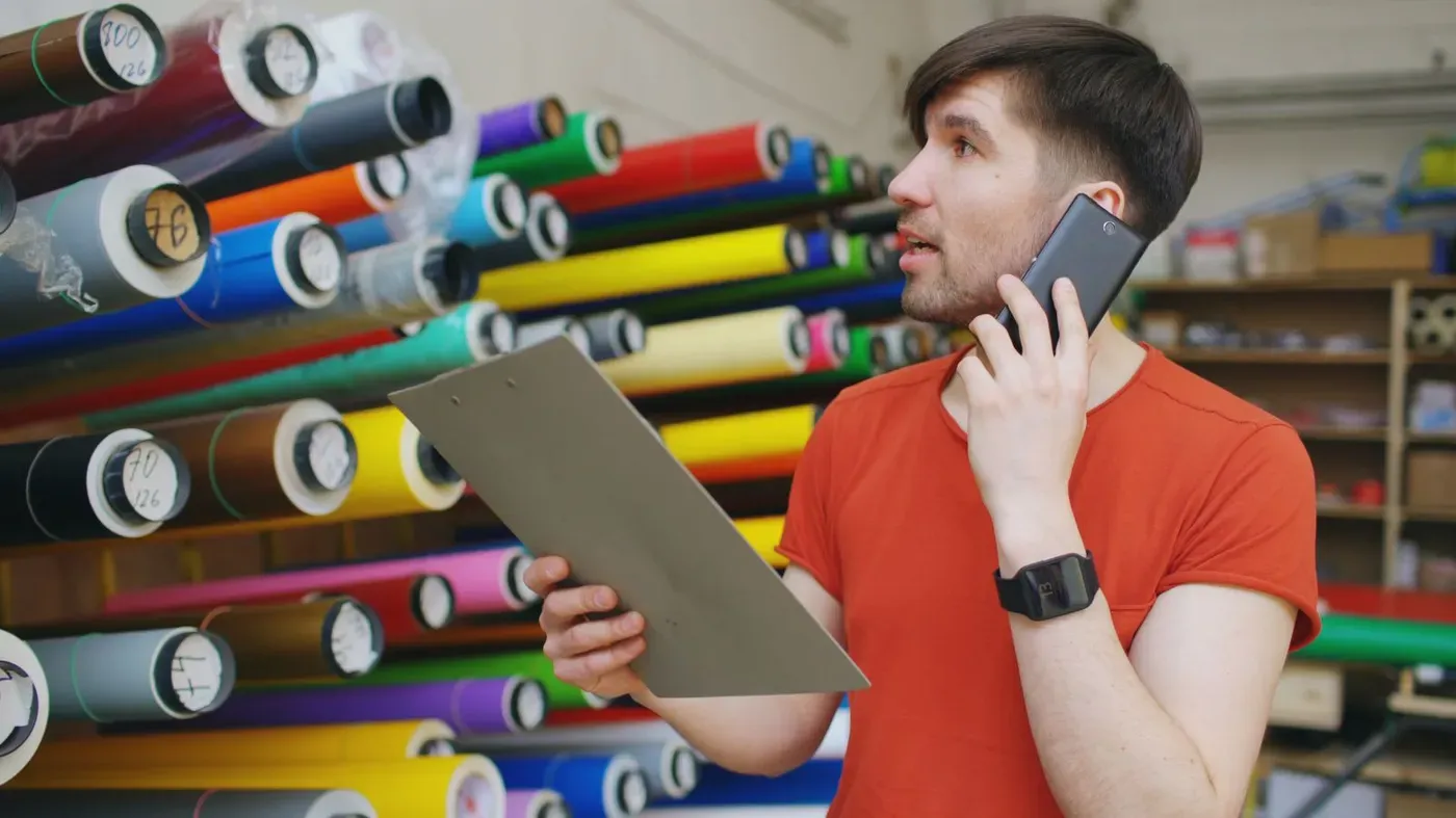 A shop owner on a phone call holding a clipboard in front of shelves of coloured material rolls.
