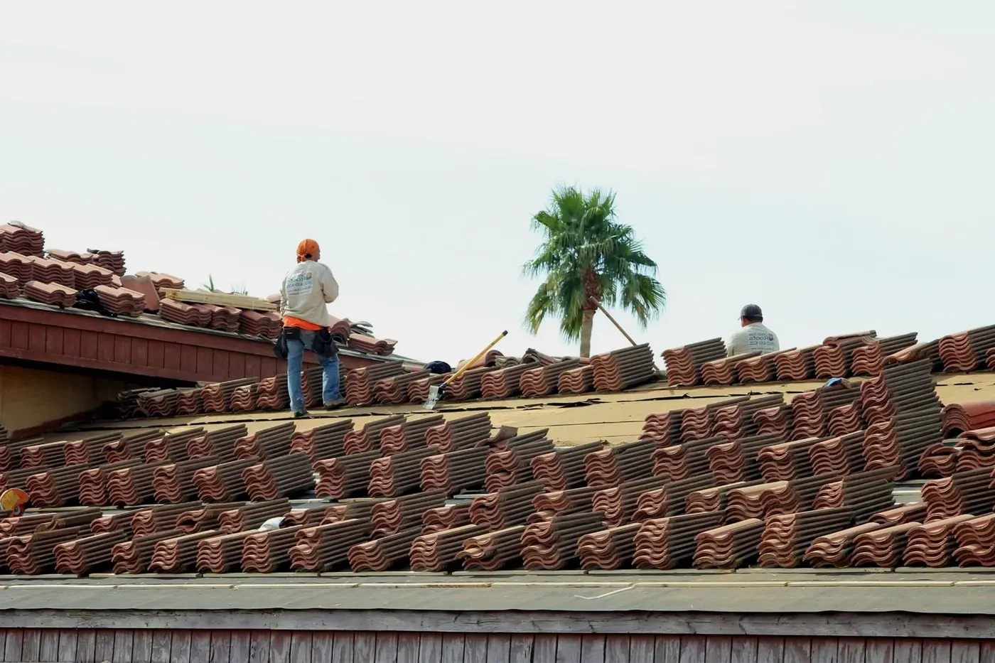 Two roofers standing on a house roof beside stacks of terracotta tiles under a clear sky.