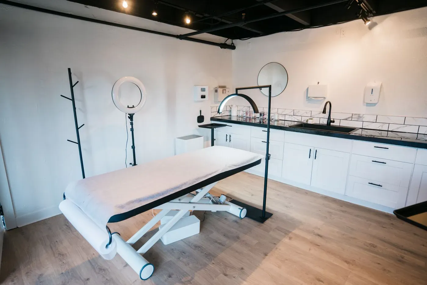 An empty treatment bed, a ring light and a sink in a white aesthetic clinic room.