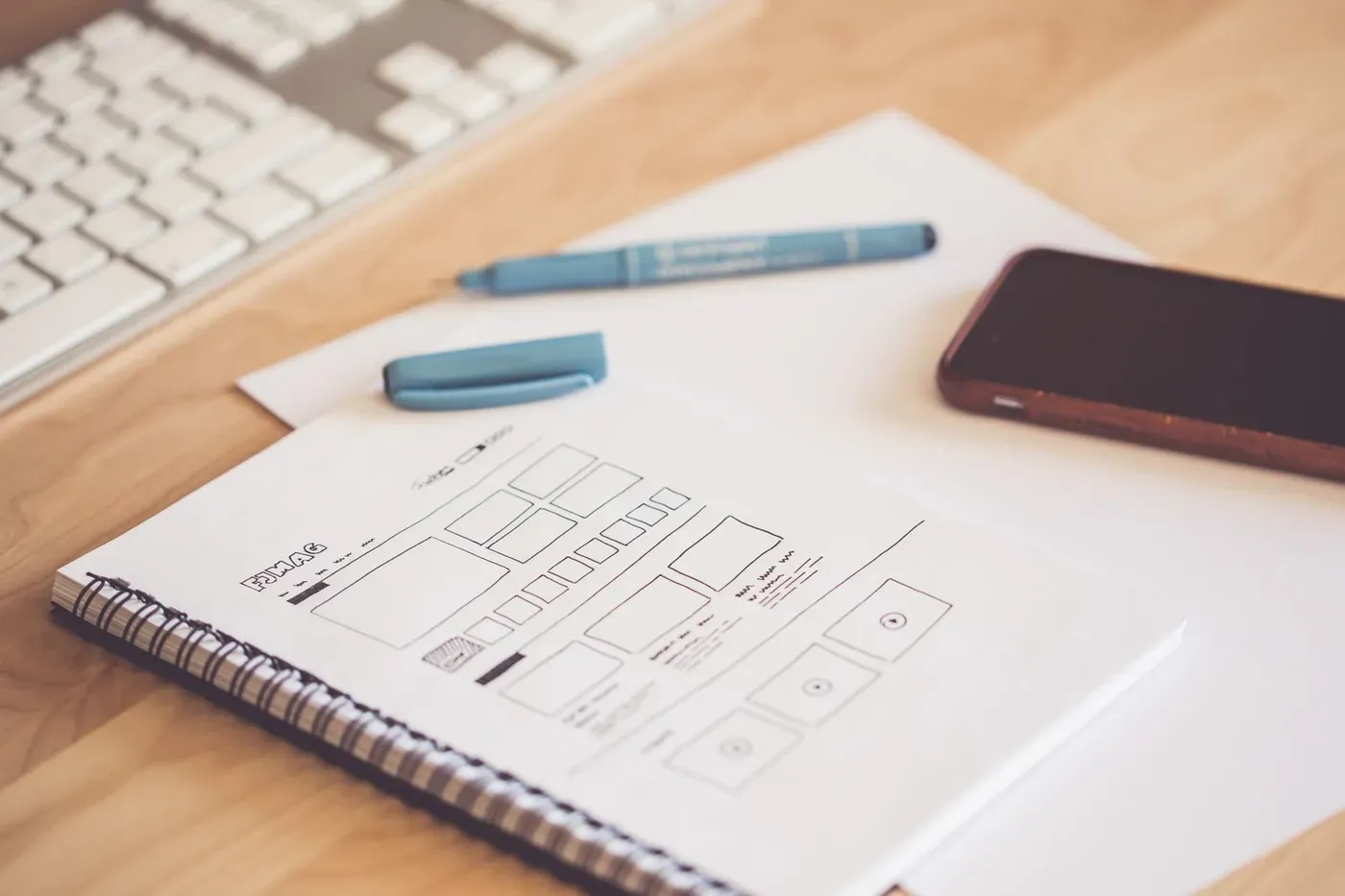 A notebook with a hand-drawn website wireframe beside a phone and a keyboard.