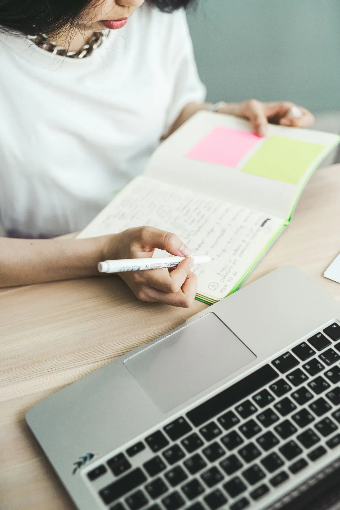 A woman writing notes in a notebook with sticky notes, next to an open laptop on a wooden desk.
